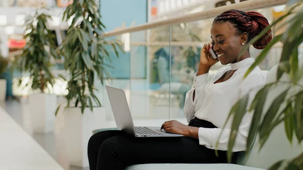 Młoda studentka Afryki siedzi na kanapie w biurze w centrum handlowym w pomieszczeniach i rozmawia przez telefon komórkowy odbierając połączenie wpisując na laptopie zdalna praca i konwersacja e-learning wielozadaniowość koncepcja — Zdjęcie stockowe