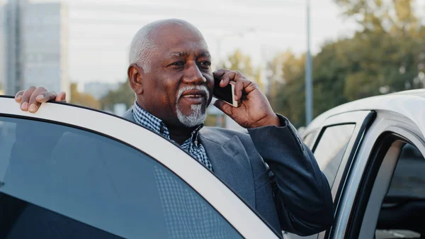 Elderly african american man talking on mobile phone walks up to car sits down in front passenger seat answers call using smartphone mature businessman discussing business conversation on telephone — 图库照片