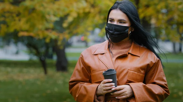 Close-up young woman stands in autumn park beautiful hispanic girl holding disposable cup of coffee wants to drink but forgets to remove medical protective mask from face shrugs up funny situation — Foto Stock