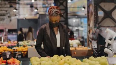 Genç Afro-Amerikalı kadın tüketici tıbbi maske takıyor. Ürünleri seçiyor. Elma meyveleri. Coronavirus pandemik alışveriş, sağlıklı yaşam tarzı diyeti.