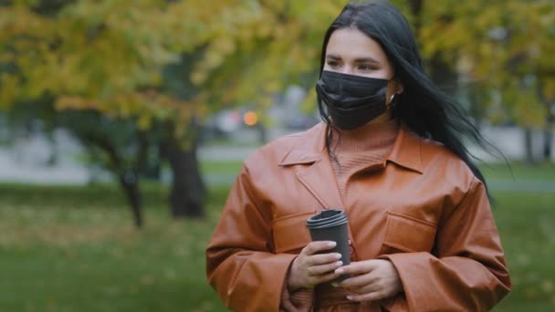 Close-up young woman stands in autumn park beautiful hispanic girl holding disposable cup of coffee wants to drink but forgets to remove medical protective mask from face shrugs up funny situation — Video Stock