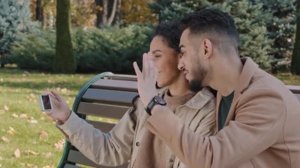 Positive hispanic couple making video call use phone man and woman smiling happily waving hello girl with boyfriend blog lovers sitting on bench in autumn park discussing outdoors talking online chat — Video Stock