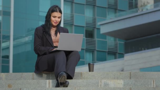 Hispanic girl sitting on steps of business center undergoing online interview using laptop presents project young businesswoman conducts videoconference on webcam distance learning via video chat — Vídeo de Stock