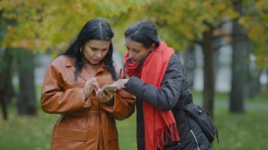 Sonbahar parkında akıllı telefon kullanan iki güzel İspanyol genç kadın internet mağazasından kıyafet seçen aletlere bakıyorlar. İnternetten yeni telefon uygulaması kullanıyorlar. Cep telefonuyla oynuyorlar.