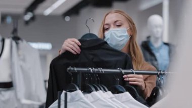 Alışveriş merkezinde maske takan beyaz bir kız elbise seçiyor. Güzel bir kadın indirimli alışveriş yapmaktan hoşlanıyor. Gömleğin kalitesini değerlendiriyor.