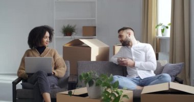 Mutlu Afro-Amerikalı kadın yeni düz dizüstü bilgisayarına bakıyor, beyaz adam akıllı telefon ekranına bakıyor, etnik eşiyle yeni bir daireye taşınıyor, çift karton kutuların yanındaki rahat kanepeye oturuyor.