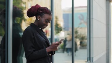 Güzel, genç, Afro-Amerikan iş kadını, kullanıcı-örnek kadın, cadde üzerinde duruyor, cep telefonuyla gülümseyen, akıllı telefon kullanarak, cihaz ile sohbet ederek mesaj yazıyor.