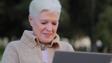 Yaşlı bayan emeklilik yaşında mutlu bir kadın dışarıda oturmuş bilgisayar kamerasına bakıyor. Çekici olgun kadın modern aletler kullanıyor. Kablosuz internet. Parkta iyi haberler var.