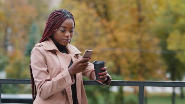 Çekici genç Afrikalı Amerikalı kız öğrenci sonbahar günü yalnız başına dışarıda kahve içiyor. Uzun saçlı milenyum kadını elinde cep telefonuyla sosyal medyada sohbet ederken akıllı telefon ekranına bakıyor. — Stok video