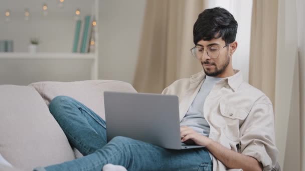 Joven árabe freelancer hombre de negocios hombre barbudo estudiante en gafas se encuentra en el sofá en casa con el ordenador portátil de trabajo estudiar lectura en línea pensando en la conexión problema frota la barbilla solución de ponderación — Vídeos de Stock