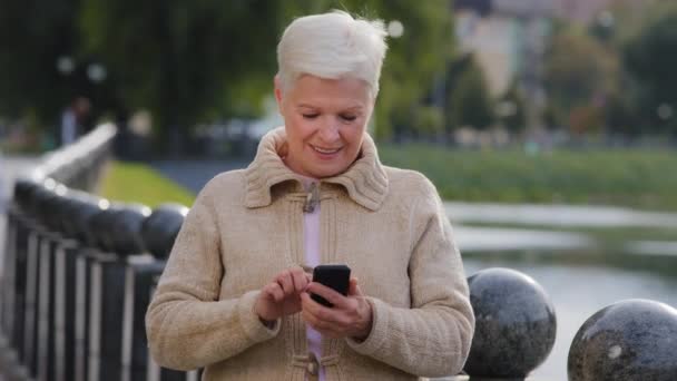 Wanita tua menarik usia pensiun menggunakan gadget modern bergulir mencari layar telepon. Betina dewasa menggunakan koneksi internet nirkabel memeriksa percakapan surat di smartphone berdiri di luar ruangan — Stok Video