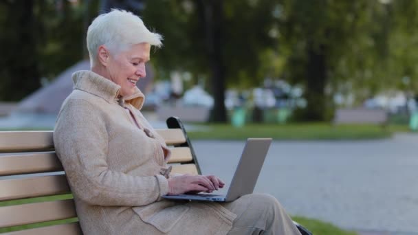 Senior gray-haired smiling woman of retirement age sitting on park bench using laptop. Granny retired typing on keyboard elderly lady browsing internet or working remotely communicating online with pc — Video Stock