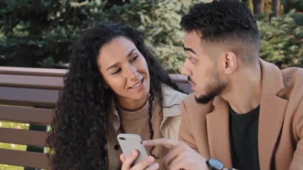 Hispanic couple sit on bench in autumn park young guy hold phone man and girl talk look at screen device surprised family smiling using app make online order male actively communicate with girlfriend — Video Stock