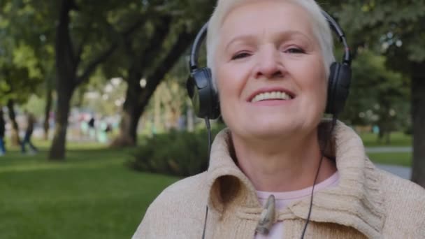 Emeklilik yaşında mutlu yaşlı bir bayan kulaklık takıyor, en sevdiği müziği dinliyor, kulaklıkla gülümsüyor, modern kulaklık takıyor, iyi niyet göstergesi olarak mutluluğu hissediyor. — Stok video