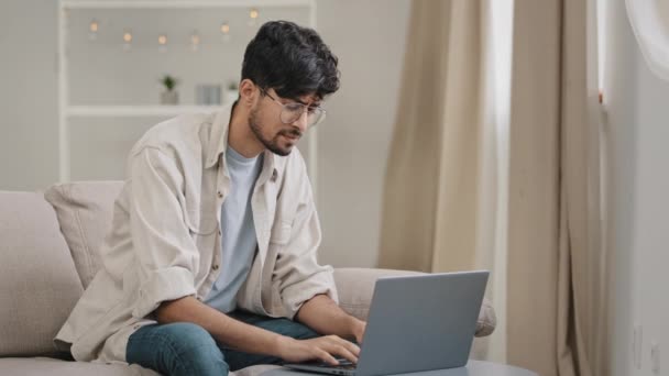 Young hispanic arab indian business man freelancer worker male bearded student wearing glasses sitting at home office on sofa with laptop feels stomach pain discomfort indigestion digestive problems — Video Stock
