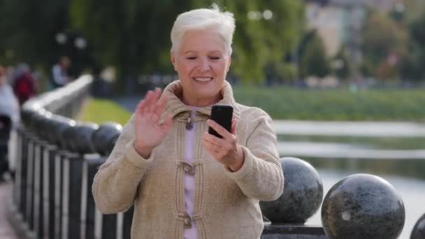 Felice donna anziana in età pensionabile fare videochiamate, guardando la fotocamera dello smartphone. Attraente vecchia donna matura utilizza gadget moderno internet wireless dire buone notizie parlando sul telefono cellulare all'aperto — Video Stock