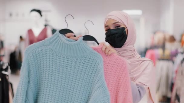 Μουσουλμάνα νεαρή γυναίκα σε ιατρική προστατευτική μάσκα κατά τη διάρκεια πανδημίας καραντίνα islamic κορίτσι σε hijab πωλητή στο κατάστημα ρούχων προσφέρει πουλόβερ προς πώληση έκπτωση θηλυκό αγοραστής δεν μπορεί να επιλέξει μεταξύ δύο χρωμάτων — Αρχείο Βίντεο