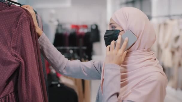 Karantina sırasındaki Müslüman kız kıyafet alışverişi sırasında kıyafet mağazasında koruyucu tıbbi maske takan İslami kadın alışverişçisi akıllı telefon kullanan seçilmiş kadın kıyafetleri giyip telefonla konuşuyor. — Stok video