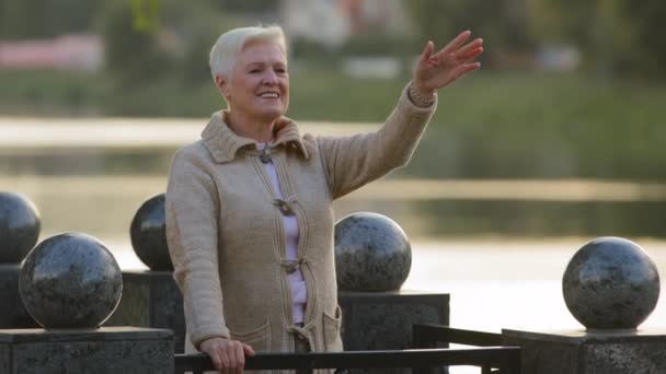 Happy middle-aged lady standing on embankment by river waves hello or goodbye, old gray-haired grandmother greets or sees off grandchildren. Elderly woman retirement age welcoming family outdoor — 图库视频影像