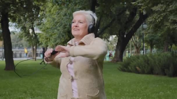 Concepto de salud de ancianos. Mujer anciana feliz y saludable, señora sonriente de la edad de jubilación usando auriculares haciendo ejercicios de calentamiento al aire libre. Abuela vieja caucásica en auriculares dispositivo digital moderno — Vídeo de stock