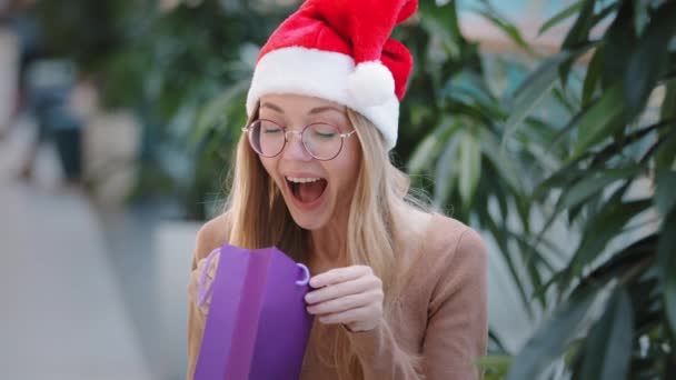 Caucasian girl in Santa hat looks in purple shopping gift bag opens cardboard package happy woman smiling rejoices buying happily dancing with present indoors millennial female joyfully celebrating — Stockvideo