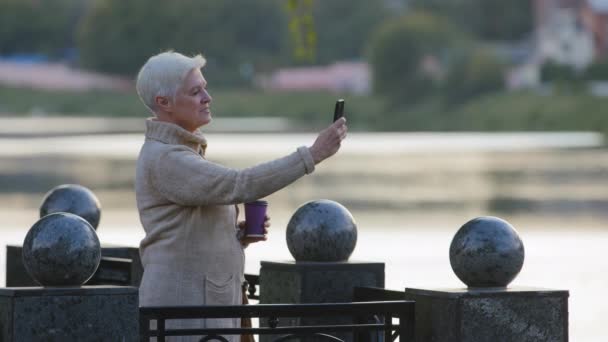 Memnun orta yaşlı bir kadın cep telefonuyla konuşan akıllı telefon ekranına bakarak video görüşmesi yapıyor. Emeklilik yaşında yaşlı bir kadın cep telefonuyla dışarıda fotoğraf çekiyor. — Stok video