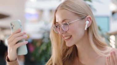 Genç milenyum kızı, alışveriş merkezinin el salında video görüşmesi yapıyor. Kablosuz kulaklıklı, mutlu, beyaz bir kadın olan arkadaşına merhaba diyor. Konuşurken eğleniyor.