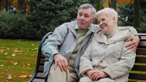 快乐的已婚夫妇笑着坐在秋天公园的长椅上年长的丈夫拥抱心爱的妻子家人在户外聊天快乐的退休人员拥抱积极的成熟配偶分享消息 — 图库视频影像