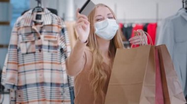 Kafkasyalı bin yıllık medikal maskeli bir kız elinde kredi kartıyla ayakta duran portre kadın indirimli alışveriş yapan mutlu alıcıyla kameraya bakıp dans eden genç bayanla eğleniyor.