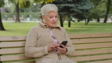 Aşırı neşeli, kır saçlı, dışarıda oturan olgun bir kadın, telefonla akıllı telefon uygulamaları kullanıyor, ekrana bakıyor, son sınıf öğrencileri ile internetten sohbet ediyor, alışveriş yapıyor, mobil cihazlarla eğleniyor.