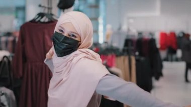 Tıbbi koruyucu maskeli mutlu genç İslamcı kadın aktif olarak müziğe taşınıyor. Müslüman kız, giyim mağazasında dans ediyor. İndirimli indirimlerden zevk alıyor.