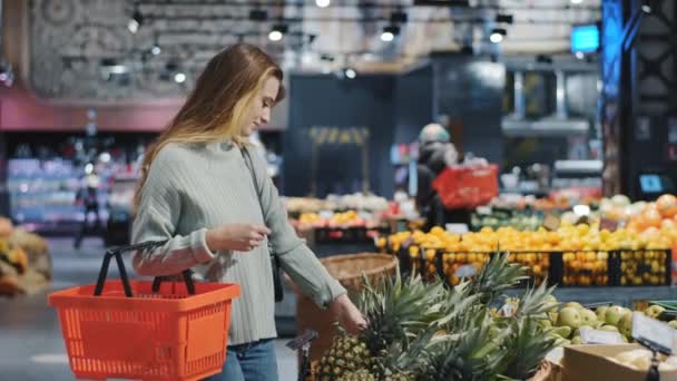 Young female shopper chooses ripe juicy healthy fruits in grocery store near counter for healthy lifestyle. Caucasian woman vegetarian with food basket buying at supermarket holding tasty pineapple — Vídeo de Stock
