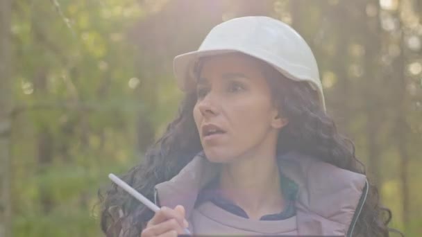 Női technikus séta, jegyzetelés vágólapon jegyzettömb papír a parkban során fakitermelés erdőirtási folyamat. Erdőértékelés és erdőgazdálkodás. Millenniumi indiai nő Erdészeti mérnök keménykalapban — Stock videók