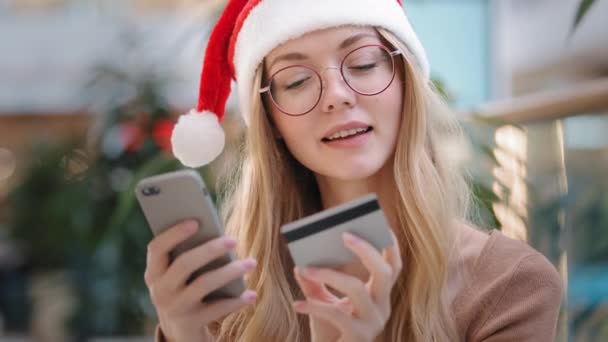 산타 모자를 쓴 캅카스 소녀는 온라인상에서 새해 선물 구매로 행복하게 웃으며 전화와 신용 카드를 사용하여 인터넷 주문을 하고 있다. — 비디오