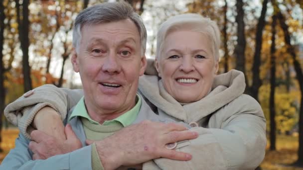 Volwassen oud vrouw knuffel geliefde man door schouders oud getrouwd paar knuffelen in de herfst park grootouders glimlachen kijken naar de camera dan elkaar genieten van communicatie buiten gelukkig huwelijk concept — Stockvideo