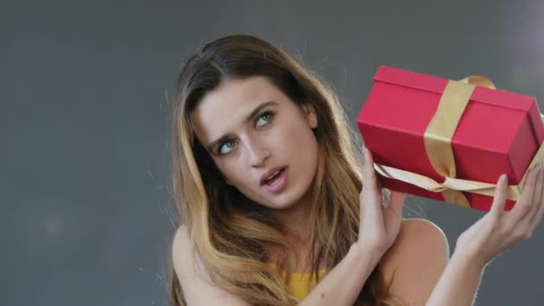 Curious young female funny caucasian lady woman model birthday girl isolated on gray studio background shakes red gift box with surprise listening present inside, congratulations holiday concept — Vídeo de Stock