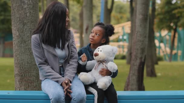 Mom and daughter sitting on bench child holding teddy bear little girl showing five fingers african american family communicates outdoors in park parent smiling listens kid mother speaking with pupil — Stockvideo