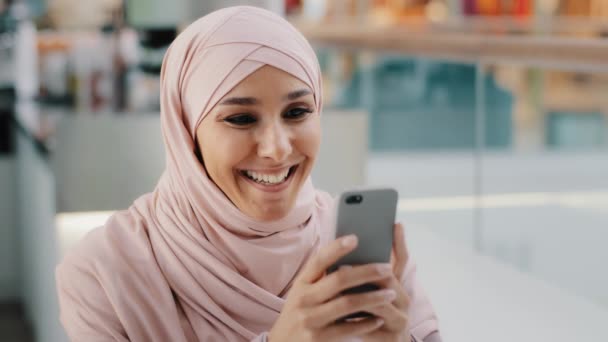 히잡 (hijab) 에 있는 행복 한 젊은 음악계 여성 이 스마트폰을 하고 있습니다. 그녀는 카페에 앉아 커피를 마시며 소셜 미디어에서 이야기를 나누며 쇼핑을 즐기고 있습니다. — 비디오