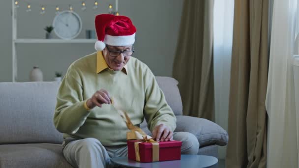 Excited elderly man in Santa hat opens box say wow show two thumb up rejoices in unexpected gift sitting on couch in home old grandfather smiling happily receiving New Year get cute Christmas surprise — Vídeo de Stock