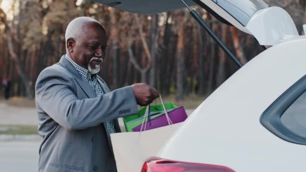 Anciano afroamericano feliz hombre pone bolsas de regalo en el coche compró regalos para las próximas vacaciones viejo hombre de negocios se regocija de compras despreocupado hombre camina hasta el automóvil cierra el maletero mirando a la cámara — Vídeos de Stock
