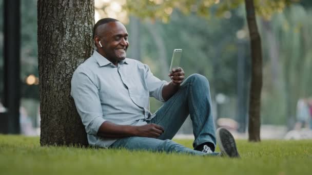Пожилой предприниматель, сидящий на траве в городском парке, бизнесмен со смартфоном, глядя на экран во время видеочата, эмоционально делится хорошей новостью с партнером — стоковое видео
