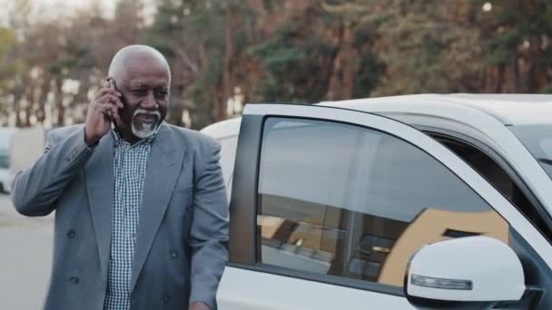 Oudere Afro-Amerikaanse man in gesprek op mobiele telefoon loopt naar de auto gaat zitten in de voorkant passagiersstoel antwoorden oproep met behulp van smartphone volwassen zakenman bespreken zakelijke gesprek over de telefoon — Stockvideo