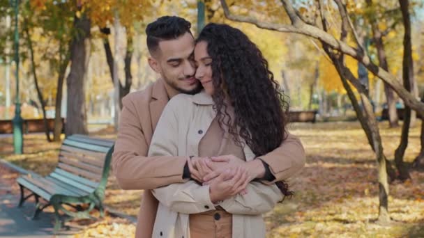 Hispanic young couple hugging standing in autumn park happy man and woman smiling enjoying date guy cuddling from back beautiful wife spouses enjoy tender moment outdoors romantic relationship concept — Stockvideo