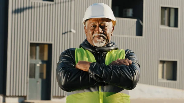 Close-up elderly african american man construction worker standing in protective helmet uniform outdoors confident workman crossed arms across chest successful builder contractor looking camera posing — стоковое фото