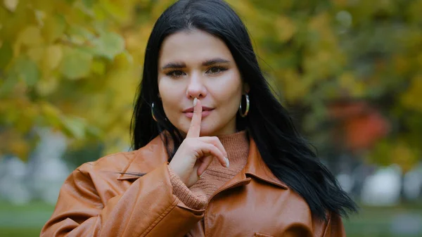 Portrait hispanic woman stands outdoors serious girl looking at camera holds index finger near mouth forbids speaking female makes gesture showing sign that asks silent about confidential information