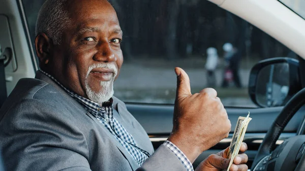 Elderly male sitting in car counting cash payment wages mature african american businessman calculates income successful business happy smiling man looking at camera shows thumbs up gesture approval — 图库照片