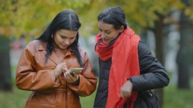 İki İspanyol kız, sonbahar parkında cep telefonu taksisi sipariş ederek sosyal ağlarda akıllı telefondan randevu sitelerinde fotoğraf izleyip online mağazadan kıyafet seçiyorlar.