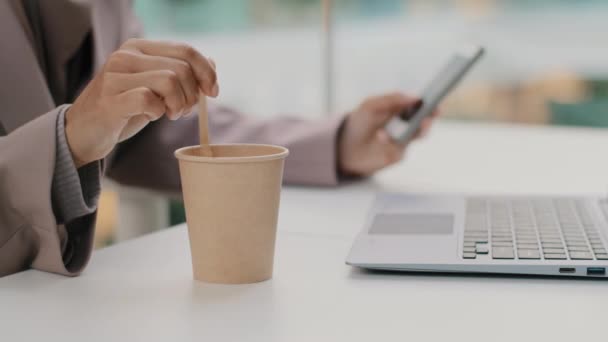 Close-up mão feminina de mulher de negócios sentada mexendo café na xícara descartável à mesa com laptop segurando telefone celular usa mensagem de digitação de aplicativo na rede social roubando fotos no smartphone — Vídeo de Stock
