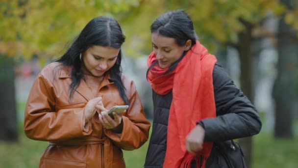 Dos niñas hispanas se paran en el parque de otoño usando mujeres telefónicas pidiendo taxi de comida en aplicación móvil viendo fotos en el sitio de citas en las redes sociales en el teléfono inteligente eligiendo la ropa en la tienda en línea — Vídeo de stock