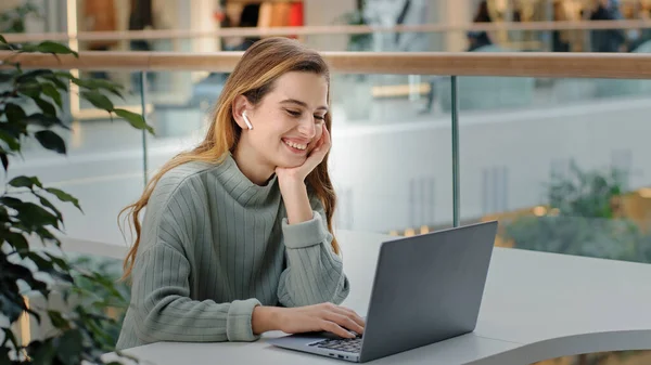 Yan bakış genç güzel bayan serbest çalışan iş kadını kablosuz kulaklıklı, alışveriş merkezinin ofisinde dizüstü bilgisayarlı, gülen dizüstü bilgisayarına bakan mutlu video mesajı okuyor.